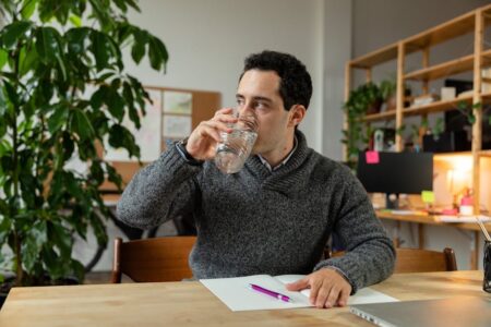 Office worker drinking water from glass, sitting at desk with plants and notes.